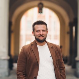 Fotografia Dawid Smułka - Uśmiechnięty mężczyzna w brązowej marynarce i białym swetrze na tle kolumnady z łukami. Portret w miejskiej scenerii, miękkie światło.