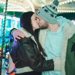 Fotografia Dawid Smułka - Zakochana para całuje się na tle karuzeli z gwiazdami. Kobieta w kurtce z futerkiem, mężczyzna w czapce z pomponem i bluzie z kapturem. Romantyczna sesja w wesołym miasteczku.