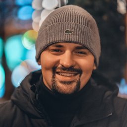 Fotografia Dawid Smułka - Uśmiechnięty mężczyzna w czarnej kurtce The North Face i szarej czapce Lacoste na tle rozmytych świateł. Portret w ciepłym oświetleniu.