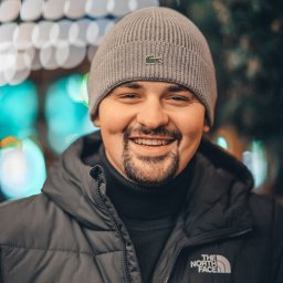 Fotografia Dawid Smułka - Uśmiechnięty mężczyzna w szarej czapce Lacoste i czarnej kurtce North Face na tle rozmytych światełek. Portret w zimowej stylizacji, ciepłe oświetlenie.