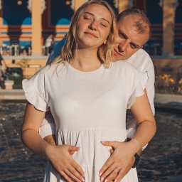 Fotografia Dawid Smułka - Ciążarna kobieta w białej sukience, obejmowana przez mężczyznę na tle fontanny i budynku z kolumnami. Romantyczna sesja w plenerze, ciepłe światło.