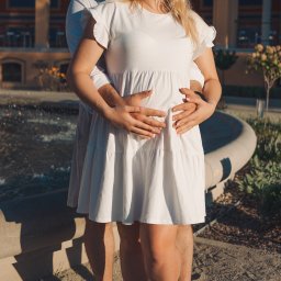 Fotografia Dawid Smułka - Czułe objęcie przyszłych rodziców w białych strojach na tle fontanny i klasycznej architektury. Dłonie mężczyzny spoczywają na brzuchu kobiety, podkreślając radosne oczekiwanie.