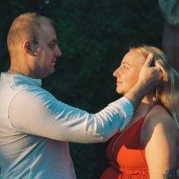 Fotografia Dawid Smułka - Czułe spojrzenie przyszłych rodziców w plenerze, kobieta w czerwonej sukience, delikatne światło podkreśla emocje, romantyczna sesja ciążowa w otoczeniu zieleni.