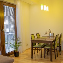 Fotografia Dawid Smułka - Jasne wnętrze jadalni z drewnianym stołem i zielonymi krzesłami, oświetlone wiszącymi lampami. Obok okna z żaluzjami stoi roślina doniczkowa.