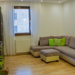 Fotografia Dawid Smułka - Jasne wnętrze pokoju dziennego z narożnikiem w odcieniach beżu i zielonymi poduszkami, drewnianą podłogą i szafką z roślinami. Okno z żaluzjami w tle.