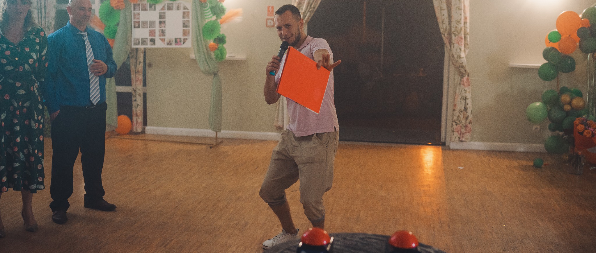 Mężczyzna z mikrofonem i pomarańczową kartką na sali balowej, dekoracje z balonów, drewniana podłoga. Prowadzący event w akcji, gestykulacja, uśmiech.