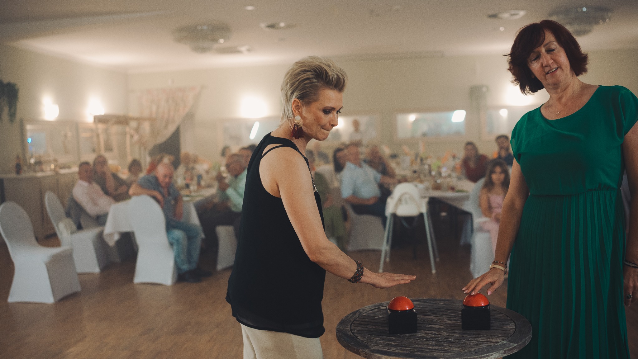 Dwie kobiety w trakcie zabawy weselnej, naciskają czerwone przyciski na drewnianym stoliku. W tle goście siedzący przy stołach, dekoracje sali.