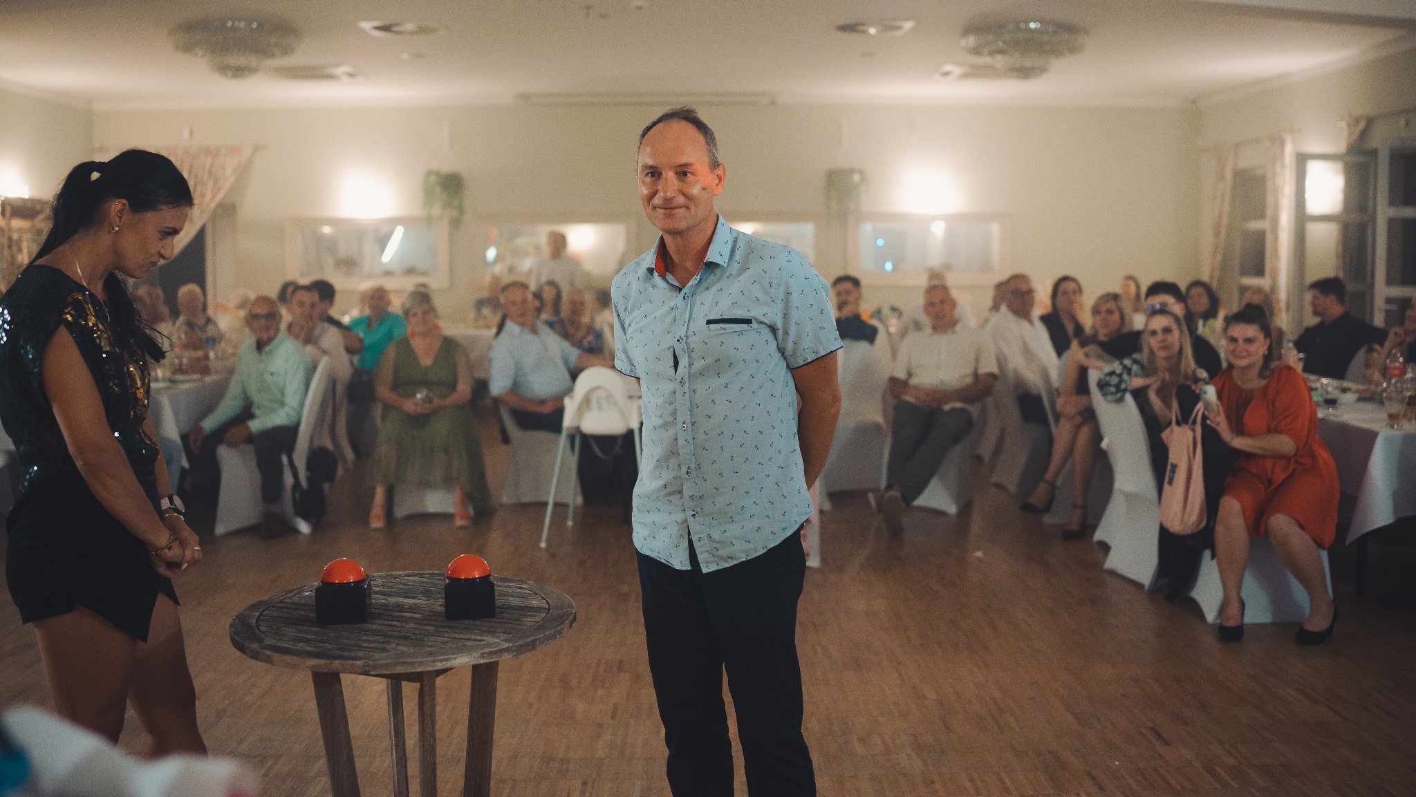 Uśmiechnięty mężczyzna na tle sali z publicznością i stolikiem z dwoma czerwonymi przyciskami, prawdopodobnie podczas konkursu lub zabawy na evencie.