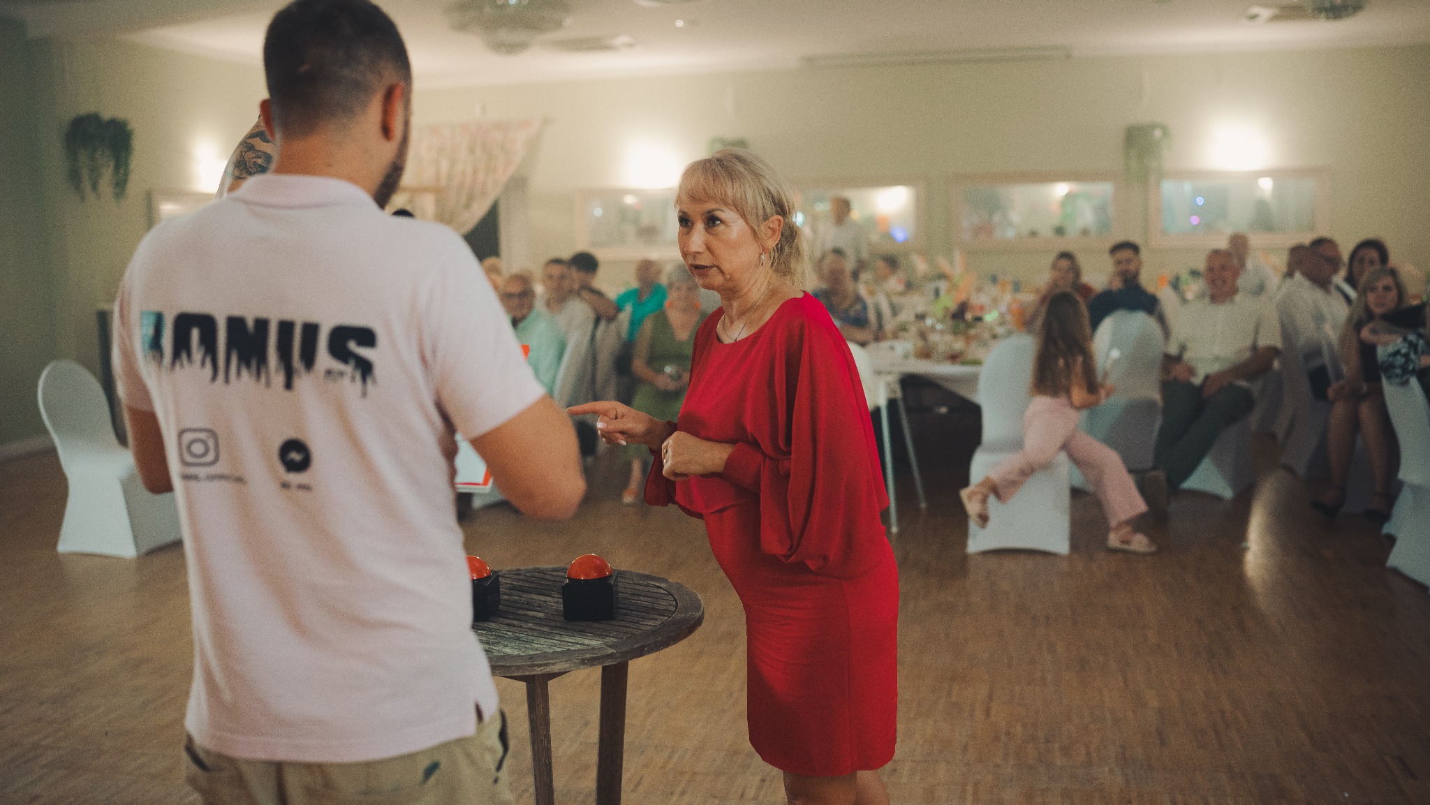 Kobieta w czerwonej sukience gestykuluje w stronę mężczyzny z tatuażem na ramieniu podczas eventu. W tle goście siedzą przy stołach.