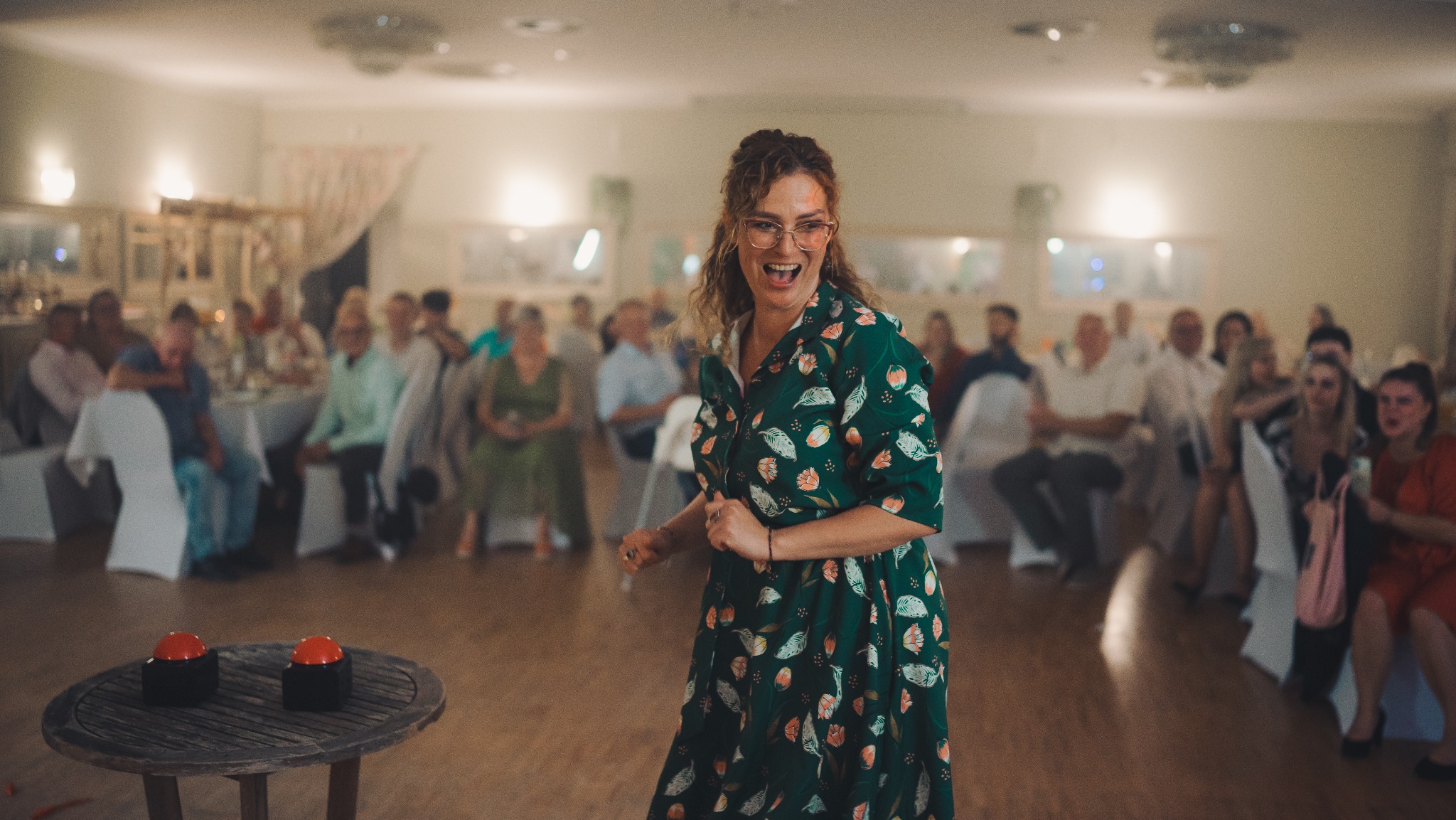 Rozbawiona kobieta w kwiecistej sukience, w okularach, tańczy na tle gości weselnych. Na pierwszym planie stolik z dwoma czerwonymi przyciskami.