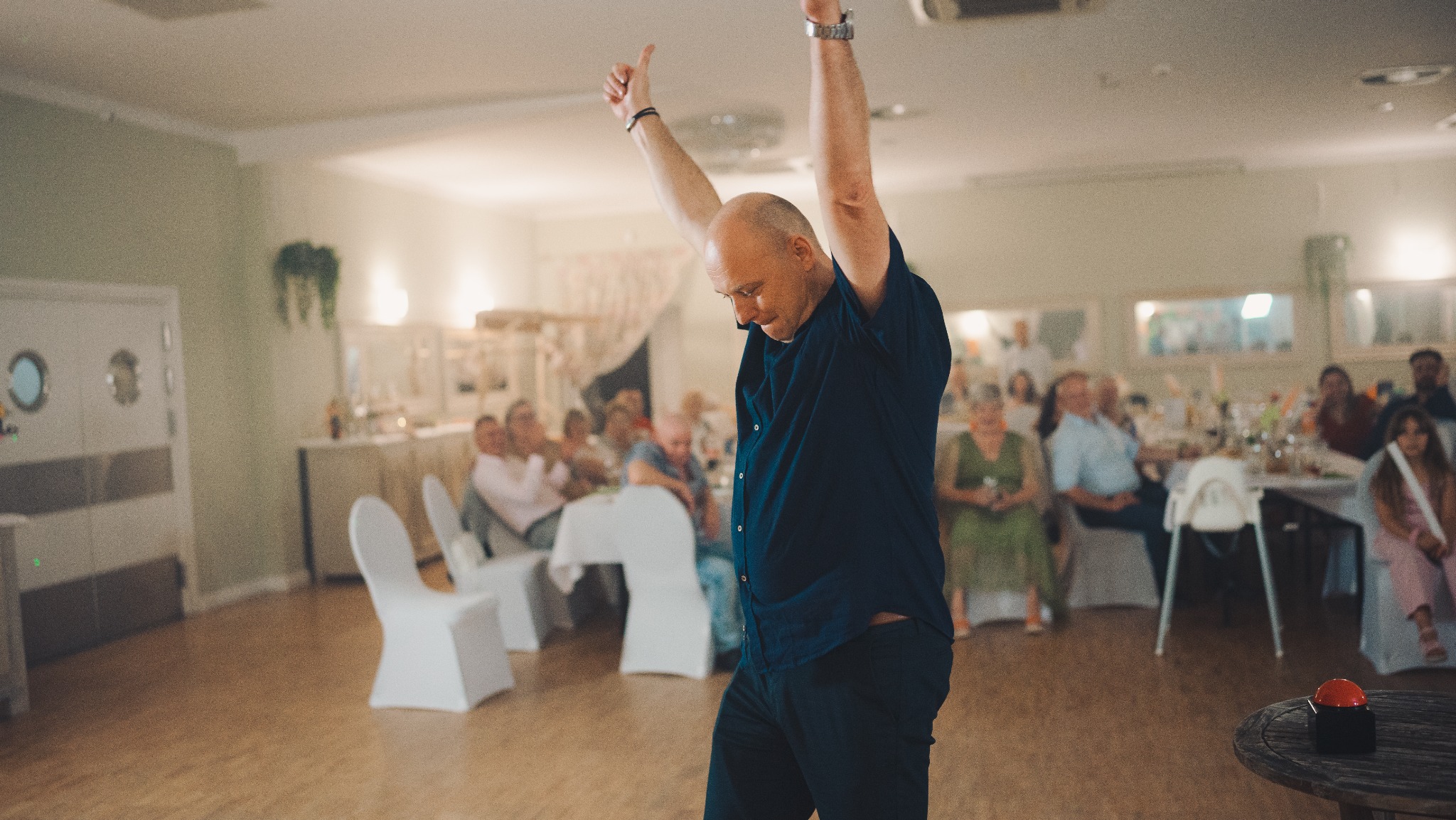Radosny mężczyzna w ciemnej koszuli tańczy z podniesionymi rękami na parkiecie, w tle siedzący goście przy stołach podczas uroczystości.