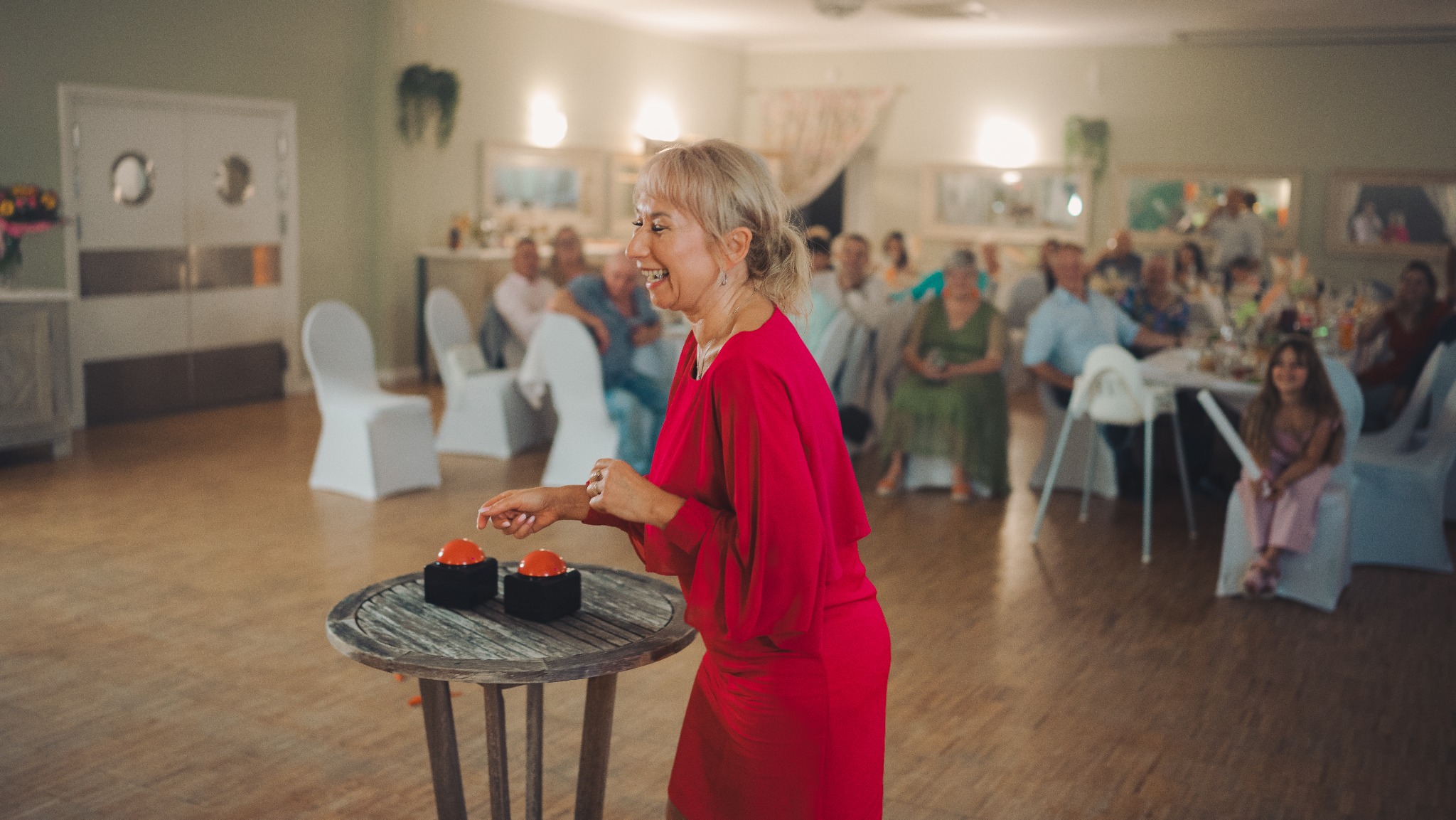 Uśmiechnięta kobieta w czerwonej sukience, w trakcie zabawy z czerwonymi przyciskami na stole, w tle goście siedzący przy stołach podczas imprezy.