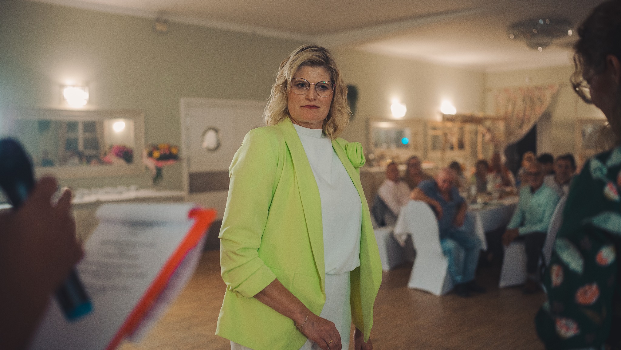 Kobieta w limonkowej marynarce, z mikrofonem i notatnikiem, przemawia na tle sali z gośćmi. Ujęcie z bliska, z widocznymi detalami stroju i mimiki.