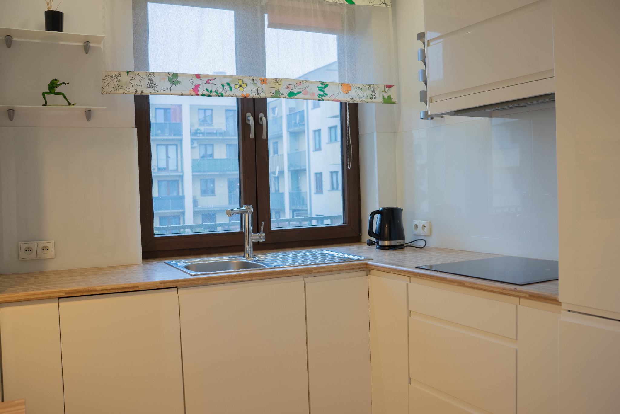 Nowoczesna, jasna kuchnia we Wrocławiu z drewnianym blatem i oknem z widokiem na budynek mieszkalny. Minimalistyczny design, czajnik elektryczny na blacie.