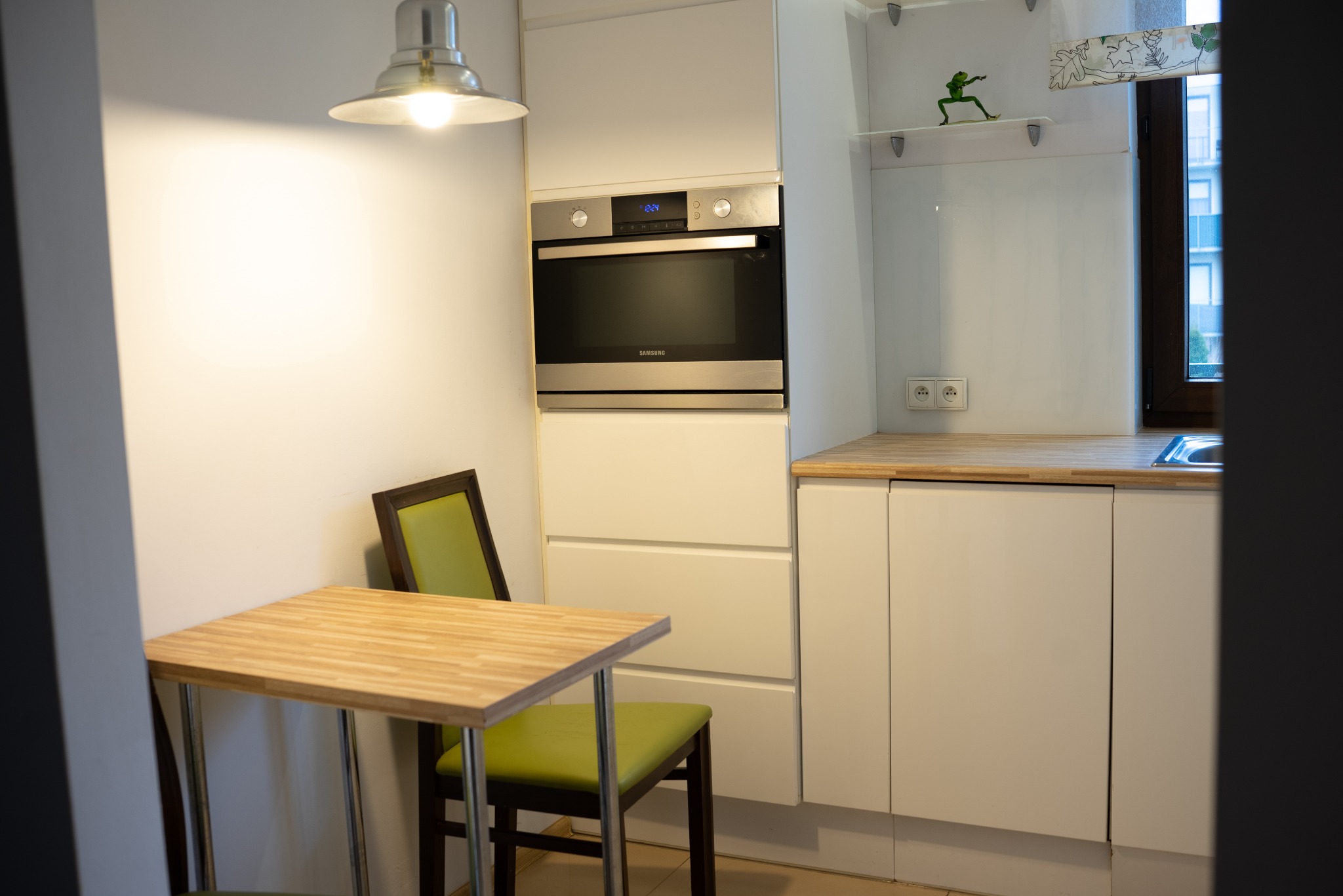 Jasna kuchnia z drewnianym blatem i zielonymi krzesłami. Na ścianie wbudowany piekarnik Samsung. Na półce figurka żaby.