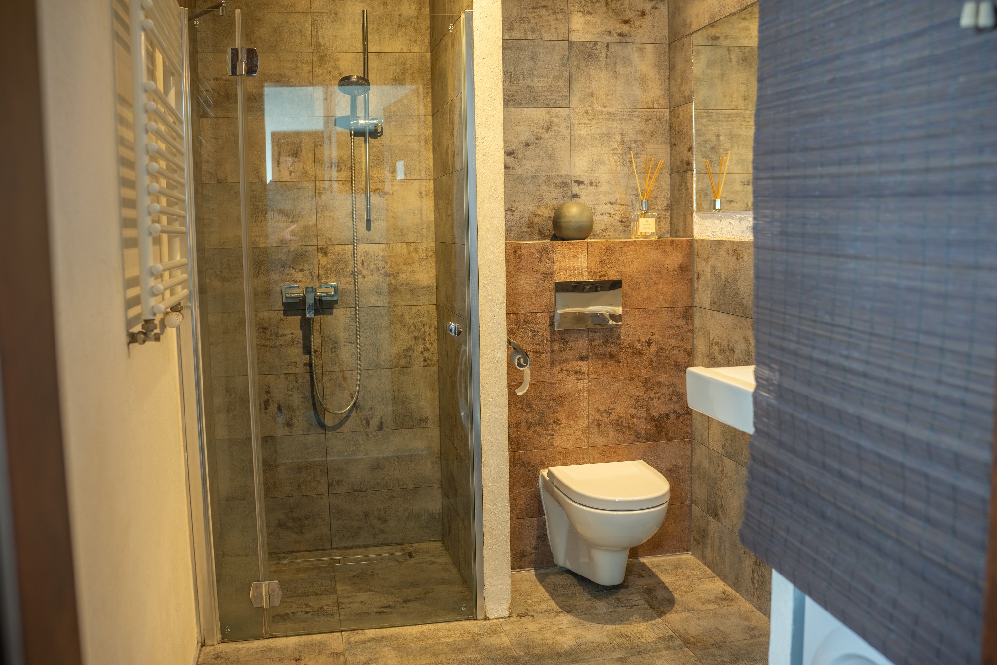 Nowoczesna łazienka z kabiną prysznicową typu walk-in, wiszącą toaletą i umywalką. Płytki w odcieniach szarości i brązu. Minimalistyczny design z akcentami dekoracyjnymi.