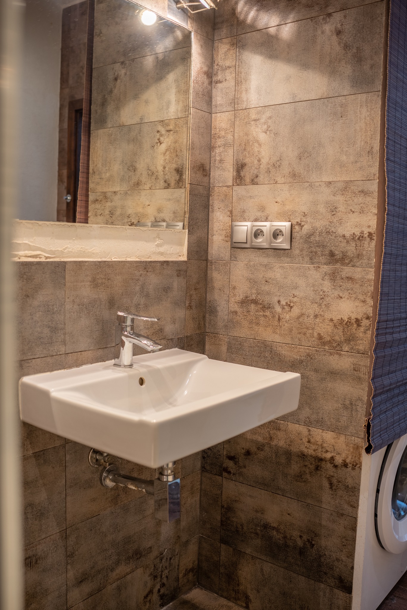 Biała umywalka z chromowaną baterią na tle płytek imitujących drewno. Nad umywalką lustro z oświetleniem i gniazdka elektryczne. W tle widoczna pralka.