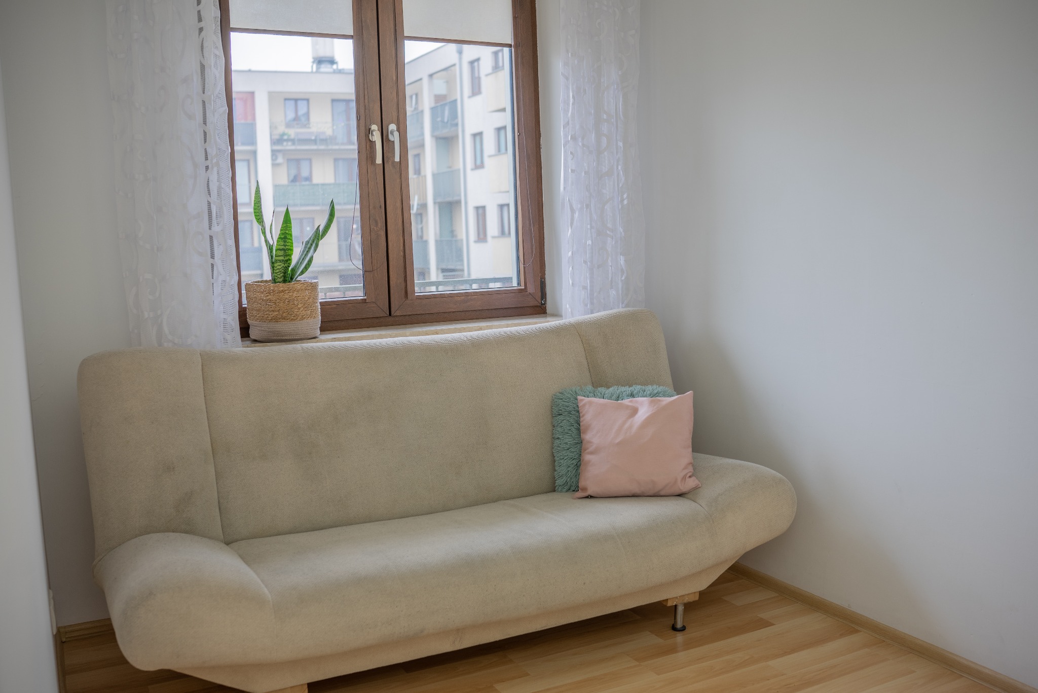 Jasna sofa z poduszkami i rośliną w wiklinowej osłonce na tle okna z widokiem na budynek mieszkalny. Minimalistyczne wnętrze w neutralnych barwach.