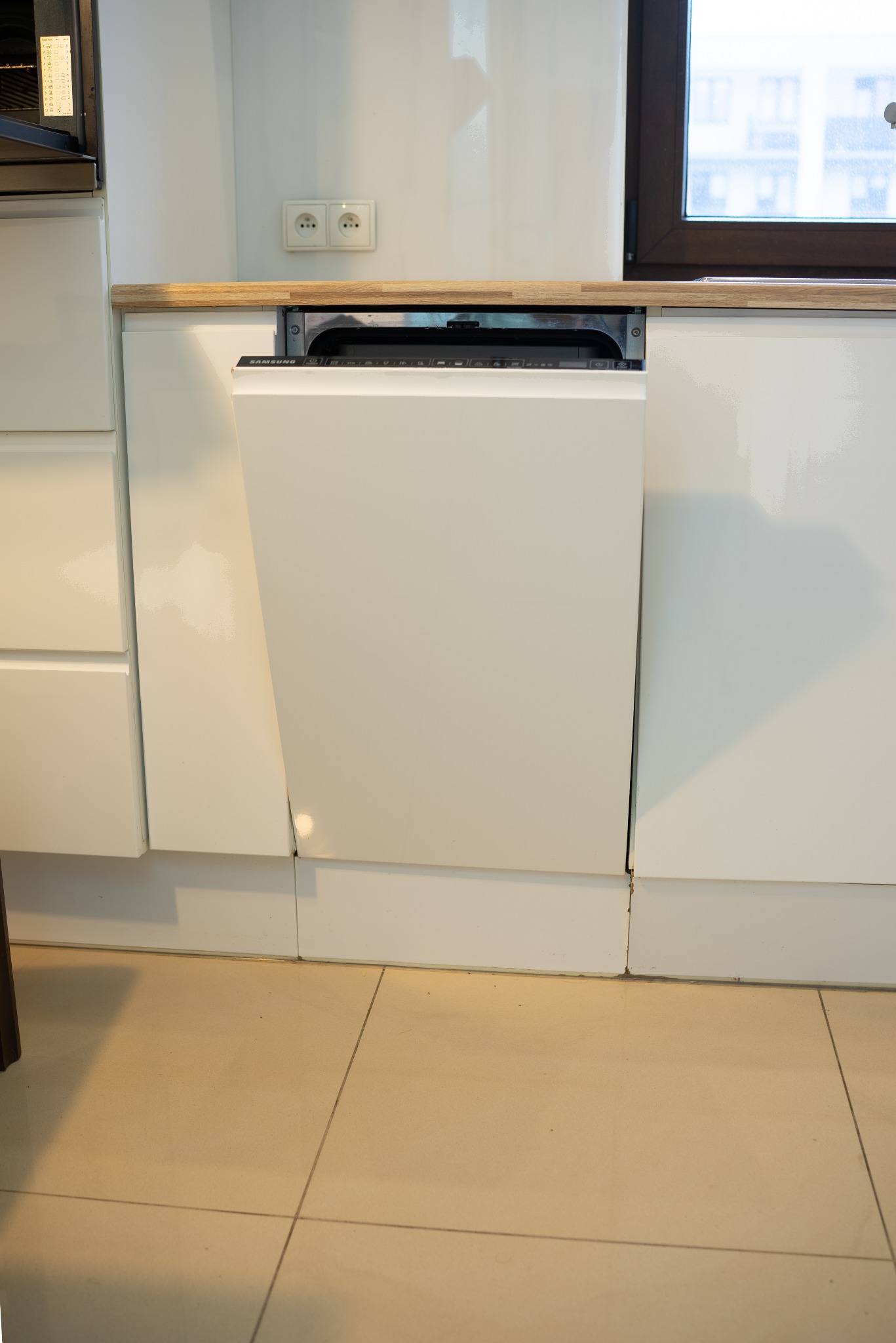Biała zmywarka Samsung w zabudowie kuchennej z drewnianym blatem, widoczna na tle jasnych płytek podłogowych i okna. Minimalistyczny design, nowoczesne wykończenie.