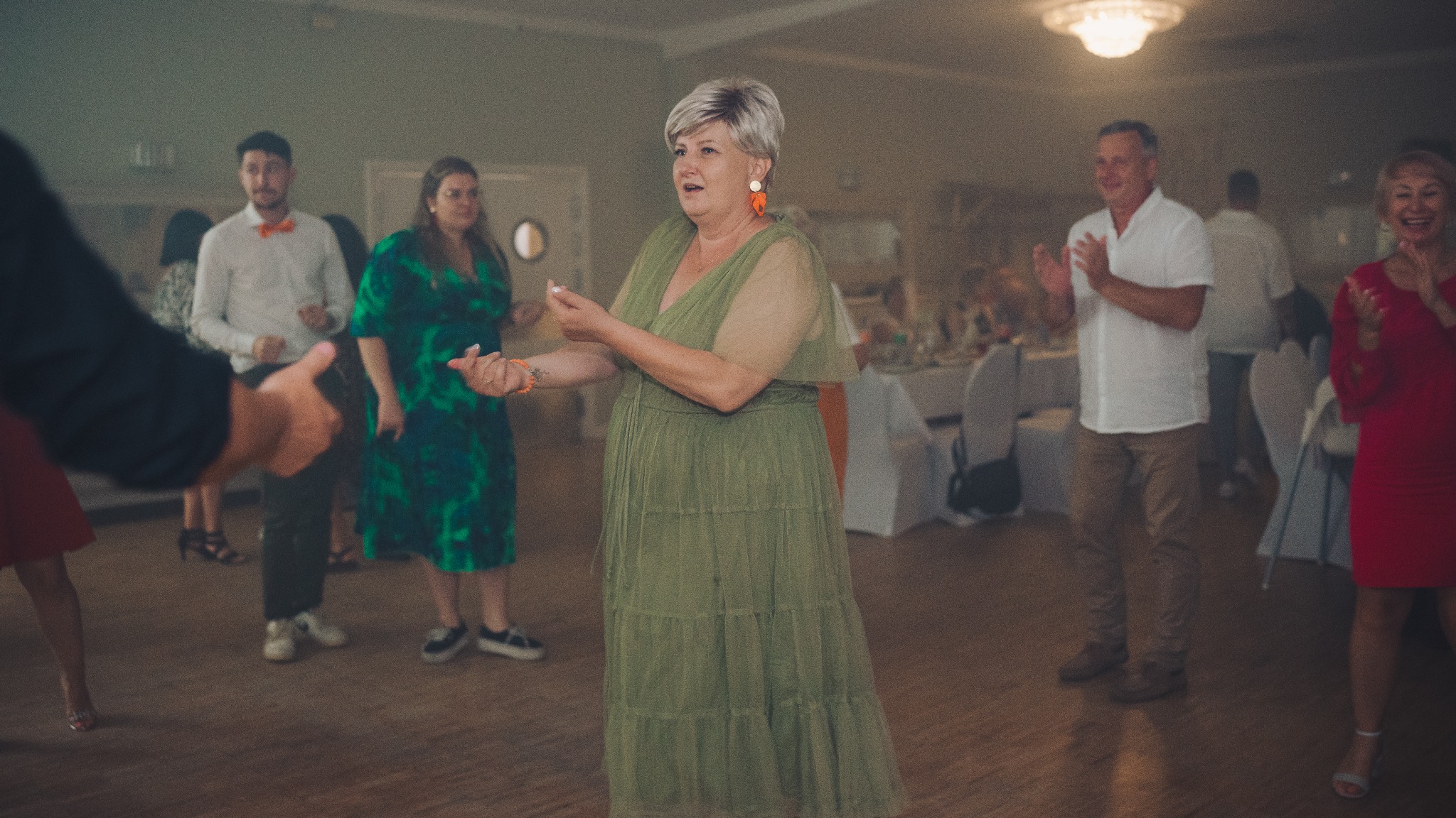 Goście weselni tańczą i klaszczą w dłonie podczas zabawy na sali. Kobieta w zielonej sukience w centrum kadru. W tle stoły i dekoracje.