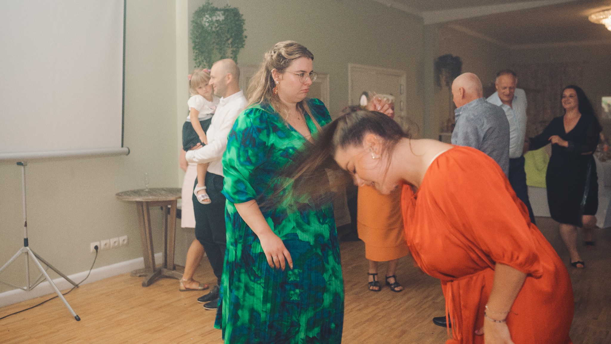 Rozmazane zdjęcie z wesela: goście w ruchu, kobieta w zielonej sukience stoi, inna w pomarańczowej odchyla głowę, w tle tańczący ludzie i rzutnik.