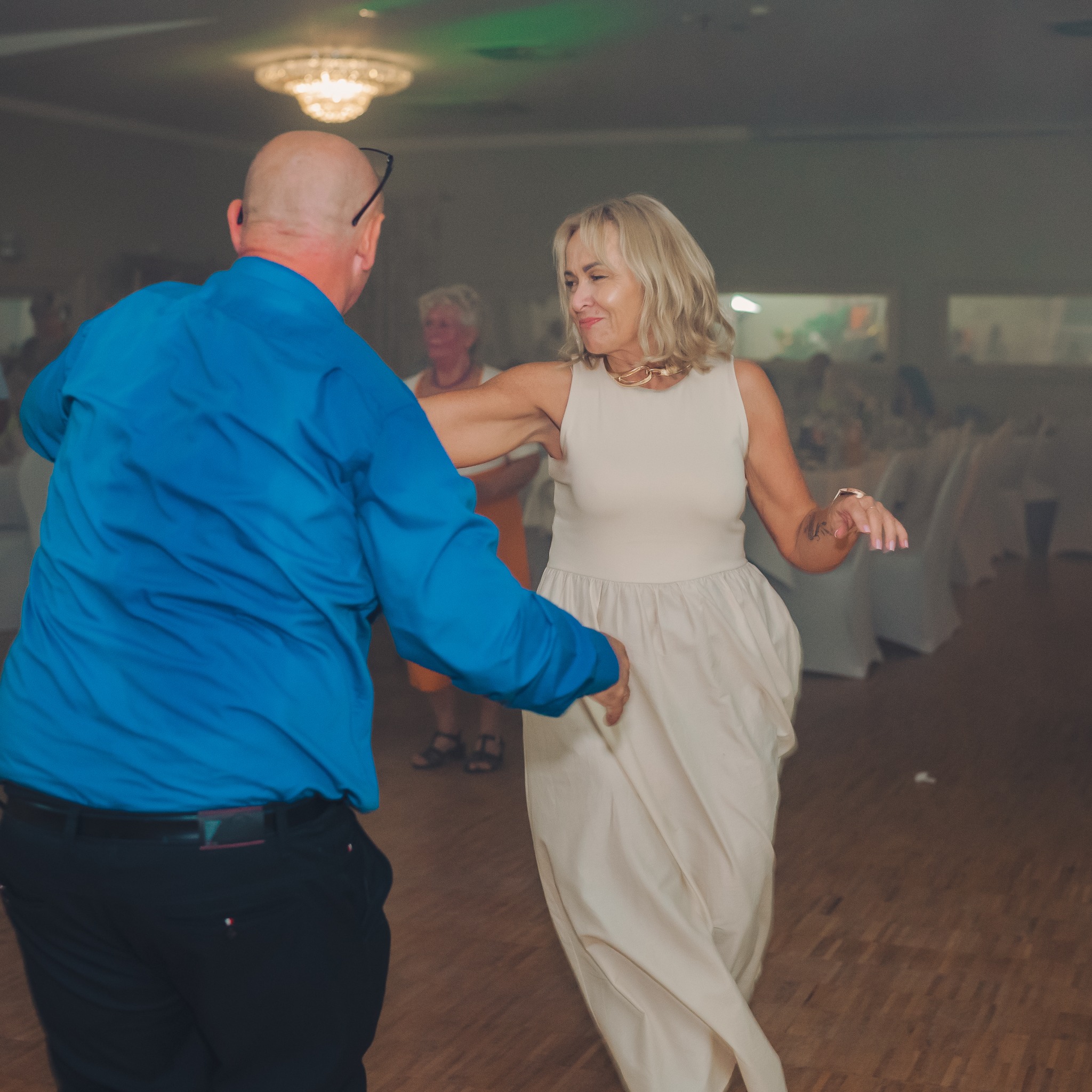 Para w tańcu podczas uroczystości, kobieta w jasnej sukni i złotej kolii, mężczyzna w niebieskiej koszuli. W tle widoczny parkiet i stoły gości.