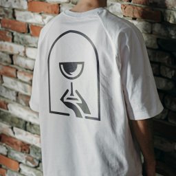 Recreate Sp. z o.o. - Biała koszulka z czarnym, minimalistycznym nadrukiem kieliszka trzymanego w dłoni na plecach, na tle ceglanej ściany. Ujęcie z tyłu, skupione na detalu grafiki.