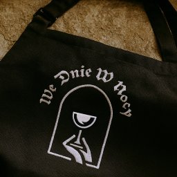 Recreate Sp. z o.o. - Czarny fartuch z białym haftem przedstawiającym kieliszek w dłoniach i napisem 'We Dnie W Nocy', leżący na tle szarej, chropowatej powierzchni.