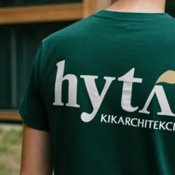 Mężczyzna w zielonej koszulce z białym logo 'hyta KIKARCHITEKCI' na plecach, stojący tyłem na tle budynku i zieleni. Logo z elementem graficznym w kolorze złotym.