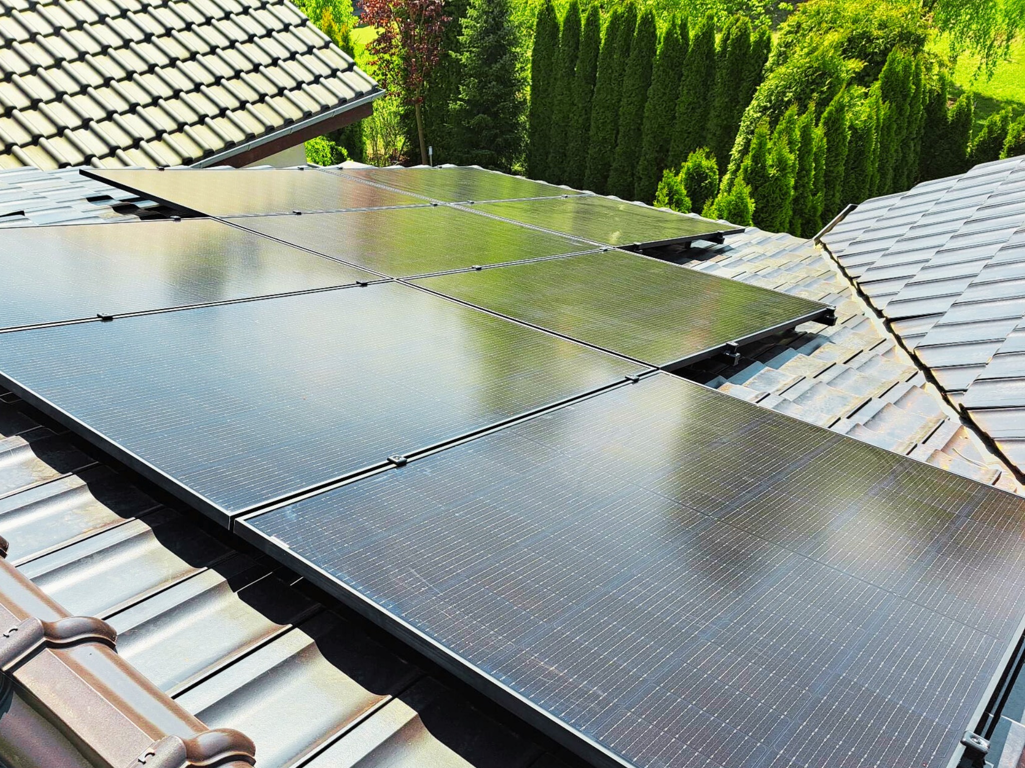 Panele fotowoltaiczne zamontowane na brązowym dachu z dachówki ceramicznej, w tle zielone drzewa.