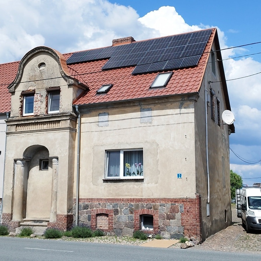 Panele słoneczne na dachu dwukondygnacyjnego domu z czerwonej cegły, widoczne okna dachowe i satelita na ścianie bocznej.