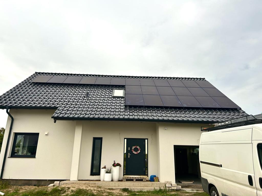 Dom jednorodzinny z zamontowanymi panelami fotowoltaicznymi na dachu pokrytym ciemną dachówką, widoczny z boku, z białym vanem zaparkowanym obok.