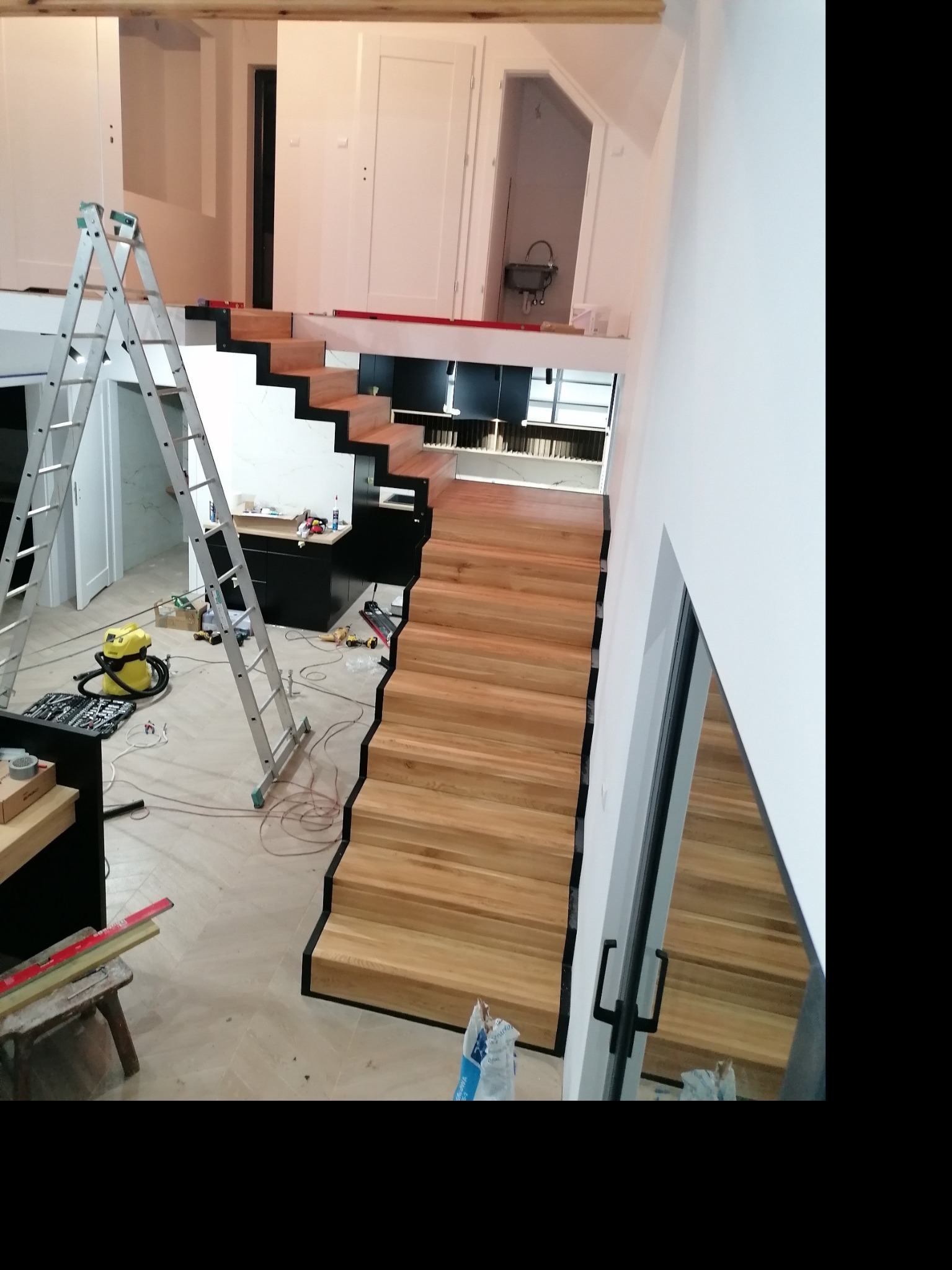 Wnętrze w trakcie wykańczania z drewnianymi schodami z czarnymi krawędziami, drabina aluminiowa, odkurzacz, narzędzia i poziomica. Widoczne także okno z odbiciem schodów.