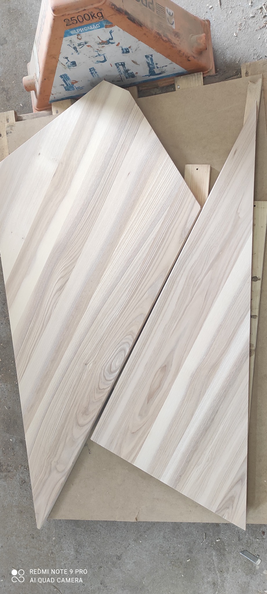 Dwa surowe, trapezoidalne elementy drewniane z wyraźnym usłojeniem, prawdopodobnie stopnie, leżące na palecie pod pomarańczowym wózkiem widłowym.