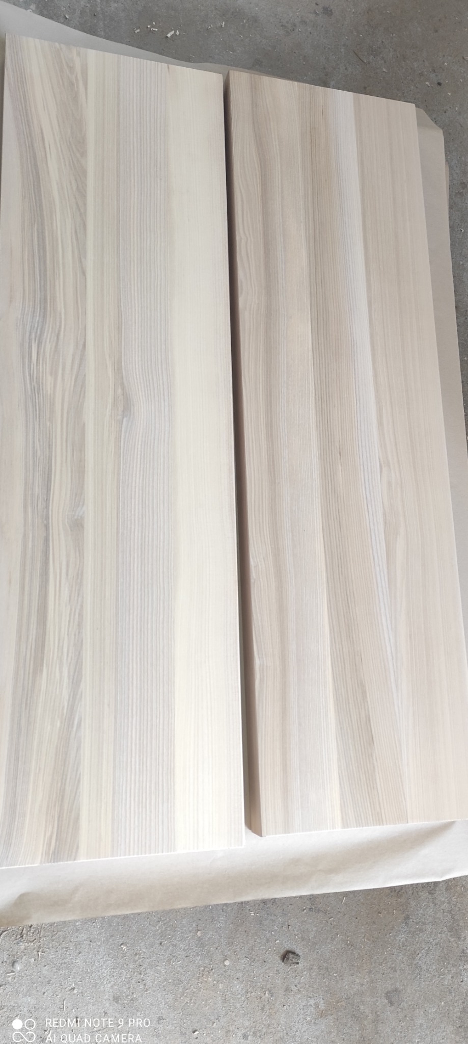 Dwie jasne, drewniane deski z widocznym usłojeniem, leżące obok siebie na szarym podłożu, prawdopodobnie w warsztacie stolarskim.