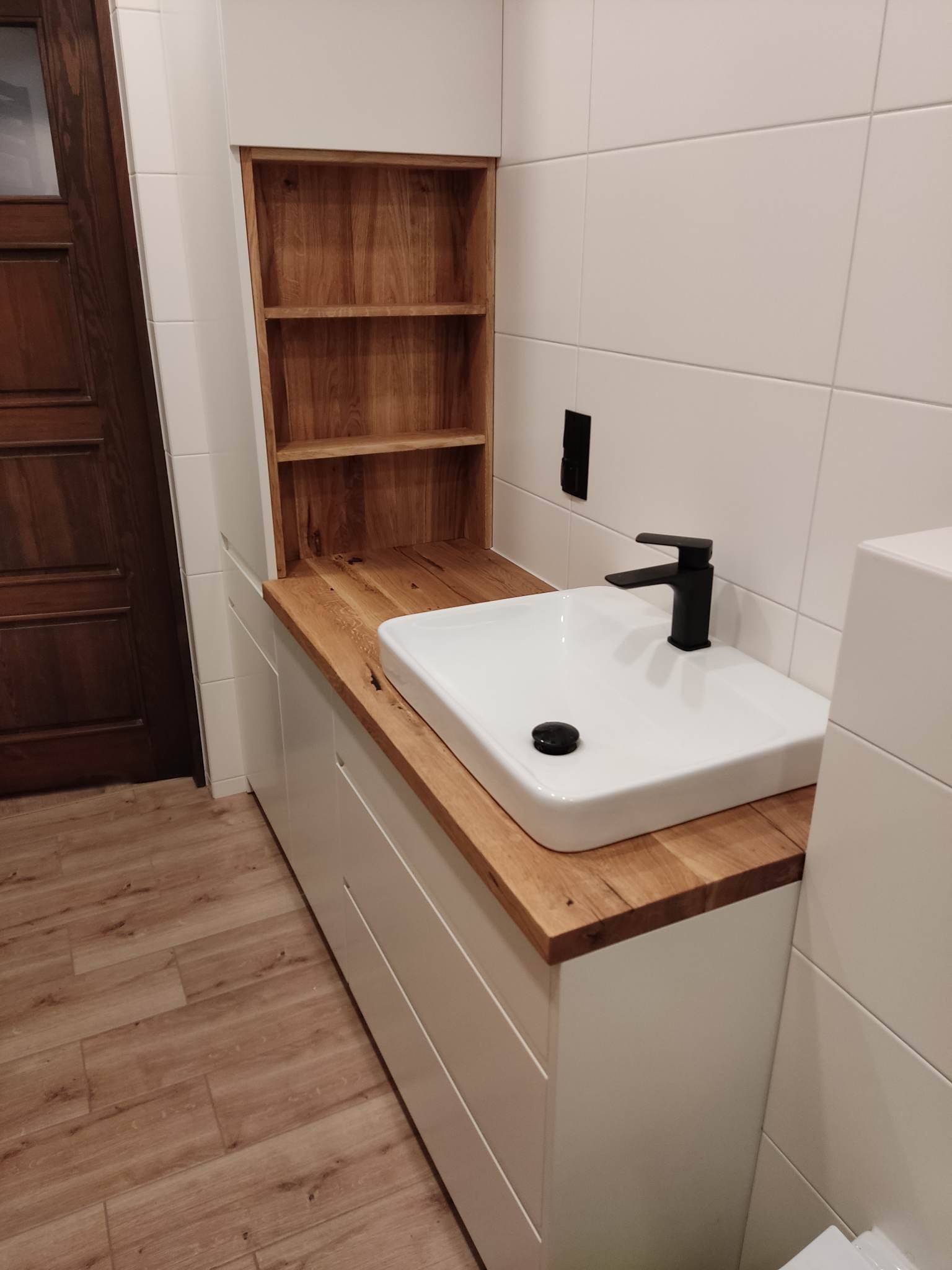 Nowoczesna łazienka z białą umywalką nablatową, czarną armaturą i blatem z jasnego drewna, uzupełniona o drewniane półki nad szafką.