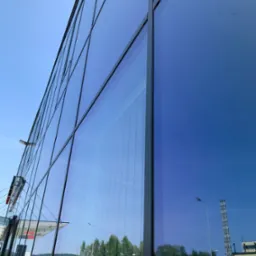 Szklana fasada budynku biurowego z widocznymi refleksami otoczenia, w tym drzew, samochodów i nieba, uchwycona pod kątem z dołu.