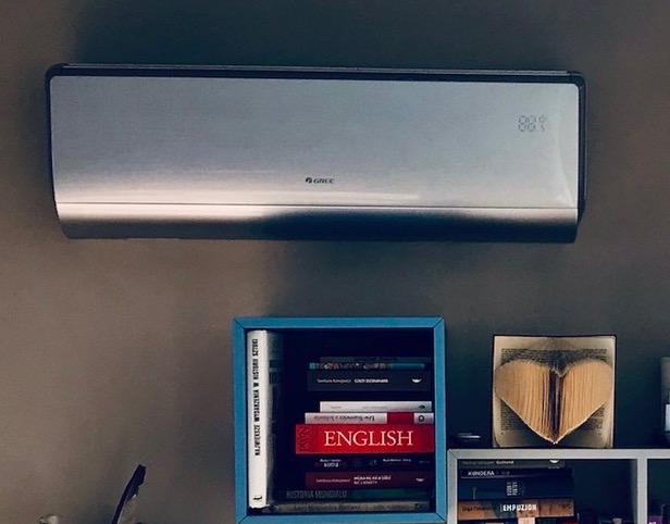 Nowoczesny klimatyzator marki Gree zamontowany na szarej ścianie nad półką z książkami, w tym z książką z napisem 'ENGLISH' i ozdobą w kształcie serca wykonaną z książki.