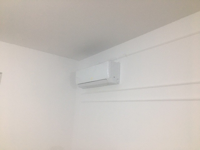 Biała jednostka klimatyzacyjna zamontowana na białej ścianie w pomieszczeniu z białym sufitem i listwami przypodłogowymi. Widok z dołu, pokazujący kabel zasilający biegnący wzdłuż ściany.