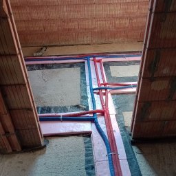 Piotr Sala Hydrosal - Montaż instalacji ogrzewania podłogowego w surowym stanie, rury grzewcze w izolacji z różowego styropianu, widoczne mocowania i fragment muru z pustaków ceramicznych.