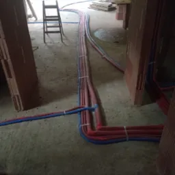 Rozprowadzenie rur instalacji wodnej i grzewczej w surowym stanie budynku z cegły, widoczna drabina i materiały budowlane w tle.