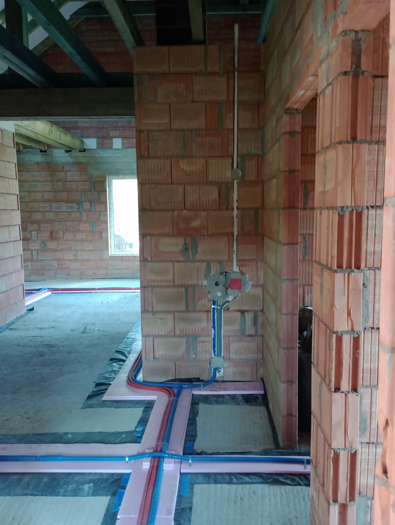 Instalacja ogrzewania podłogowego w surowym stanie, rury grzewcze ułożone na styropianie, widoczne przyłącze wodne na ścianie z cegły dziurawki, w tle okno i drewniana konstrukcja dachu.