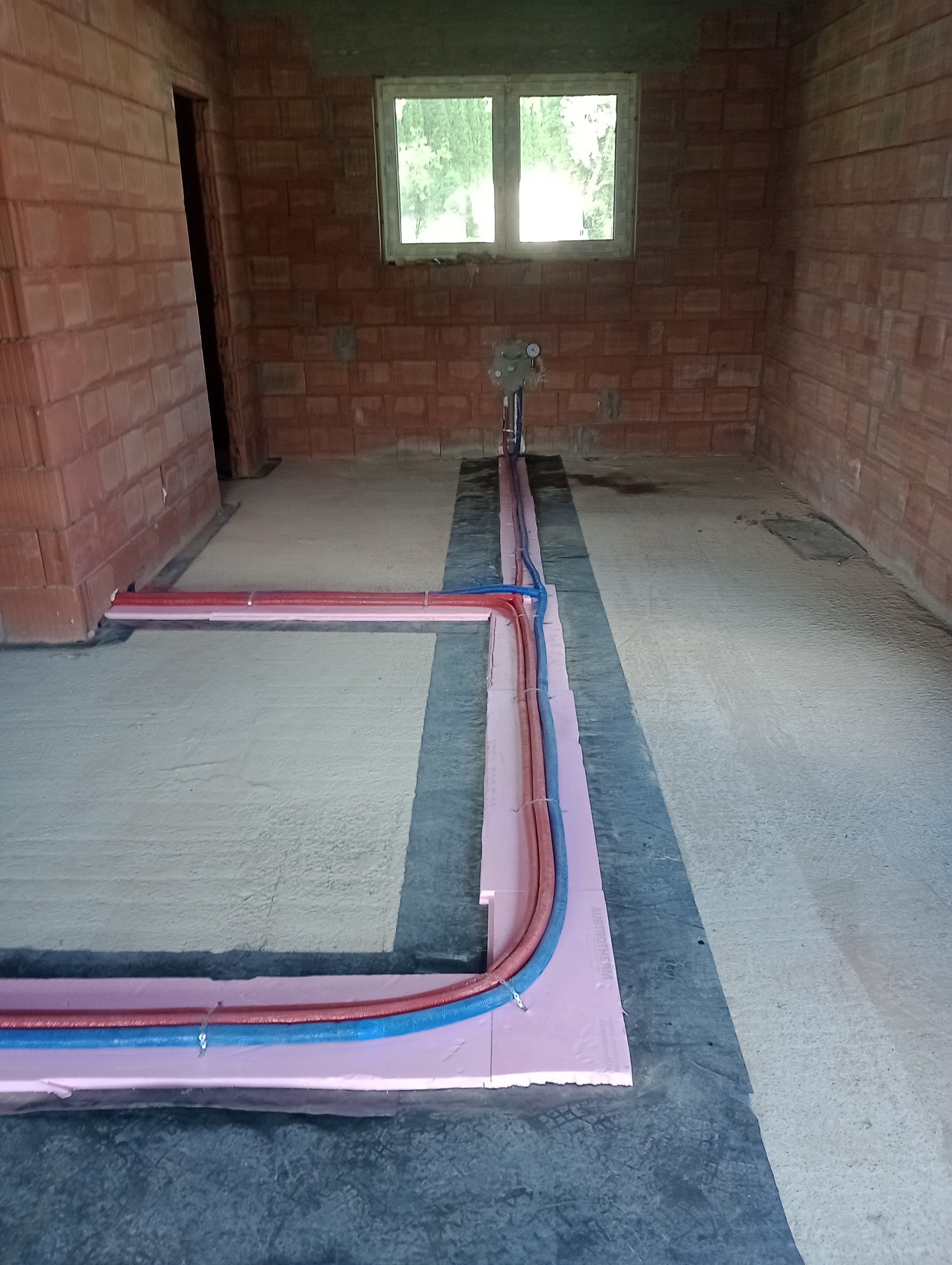 Wnętrze w trakcie budowy z widocznymi czerwonymi i niebieskimi rurami na różowym styropianie, przygotowanymi do instalacji ogrzewania podłogowego, na podłożu pokrytym czarną folią, z surowymi...