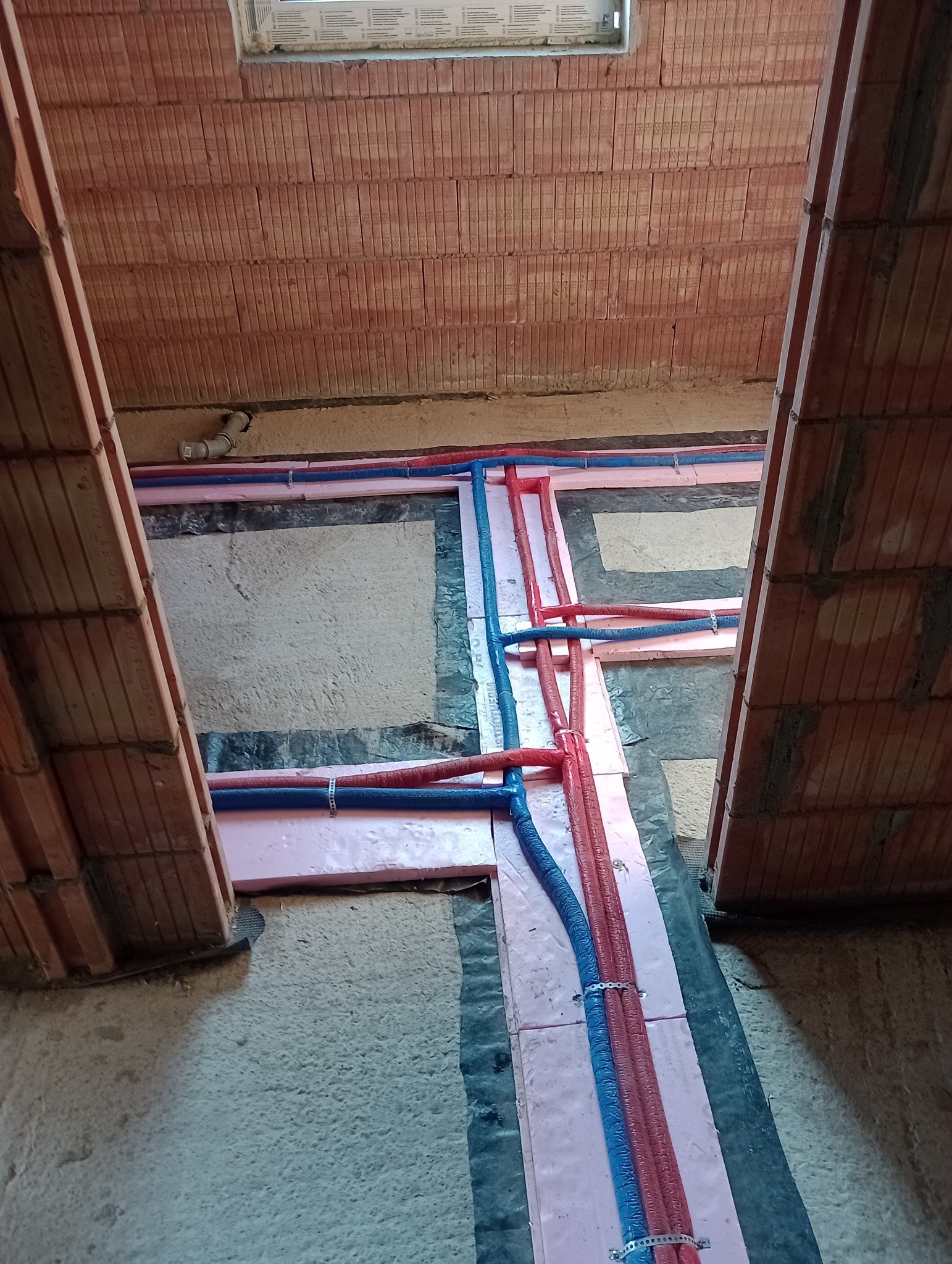 Montaż instalacji ogrzewania podłogowego w surowym stanie, rury grzewcze w izolacji z różowego styropianu, widoczne mocowania i fragment muru z pustaków ceramicznych.
