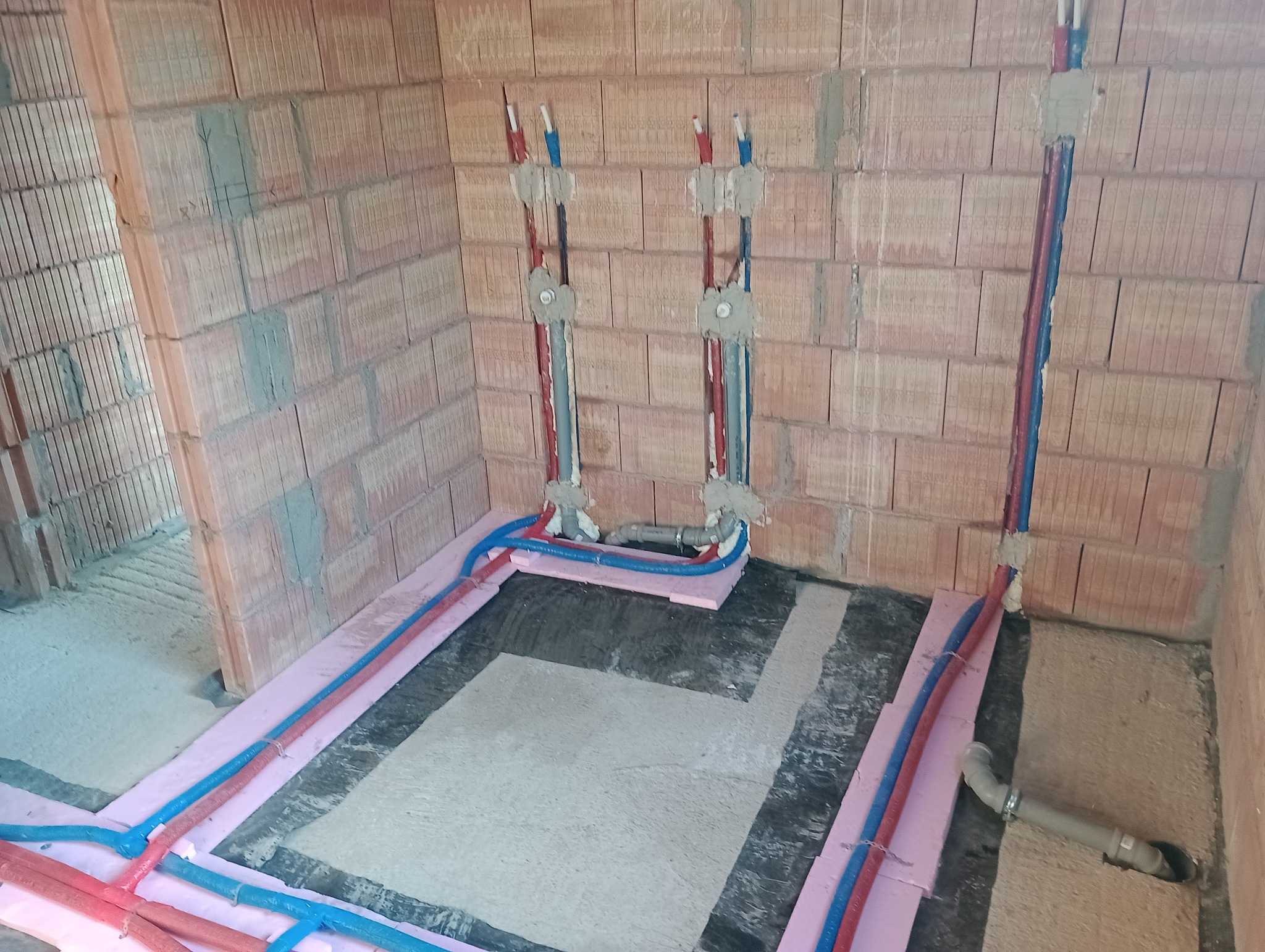 Instalacja ogrzewania podłogowego i przyłącza wodno-kanalizacyjnego w surowym stanie, widoczne rury PEX w izolacji termicznej oraz podejścia wod-kan w ścianie z cegły.