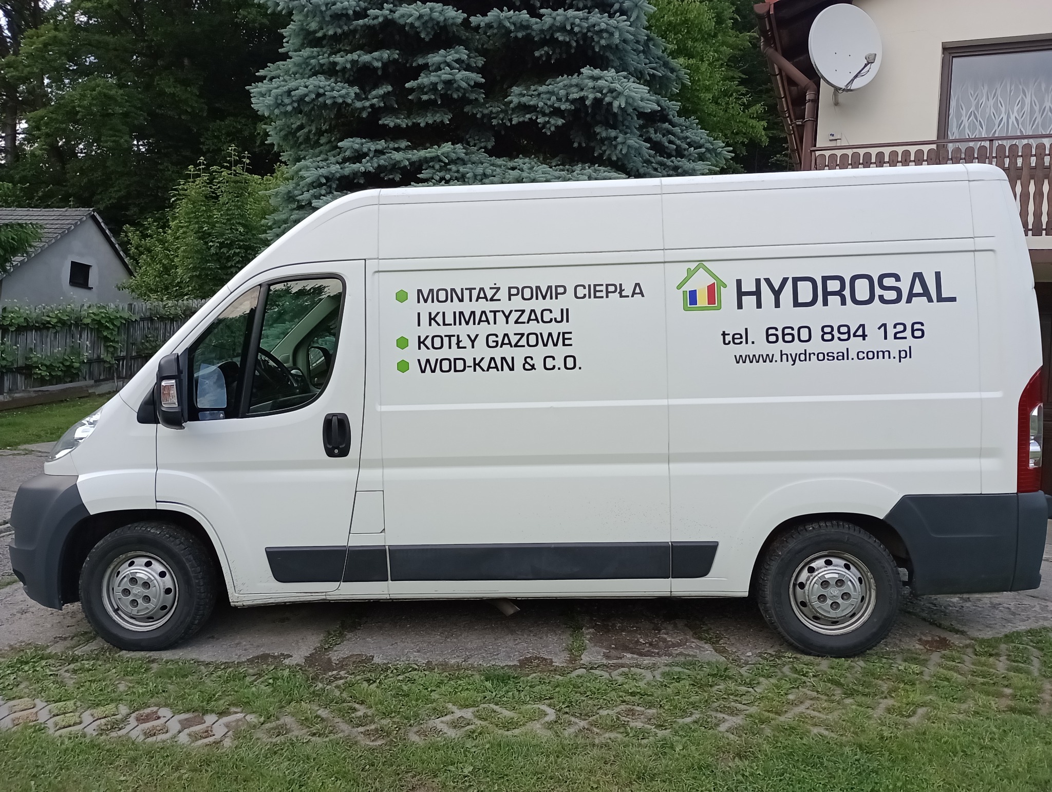 Biały samochód dostawczy firmy Hydrosal z reklamą montażu pomp ciepła, klimatyzacji, kotłów gazowych i instalacji wod-kan, zaparkowany na trawie przed domem.