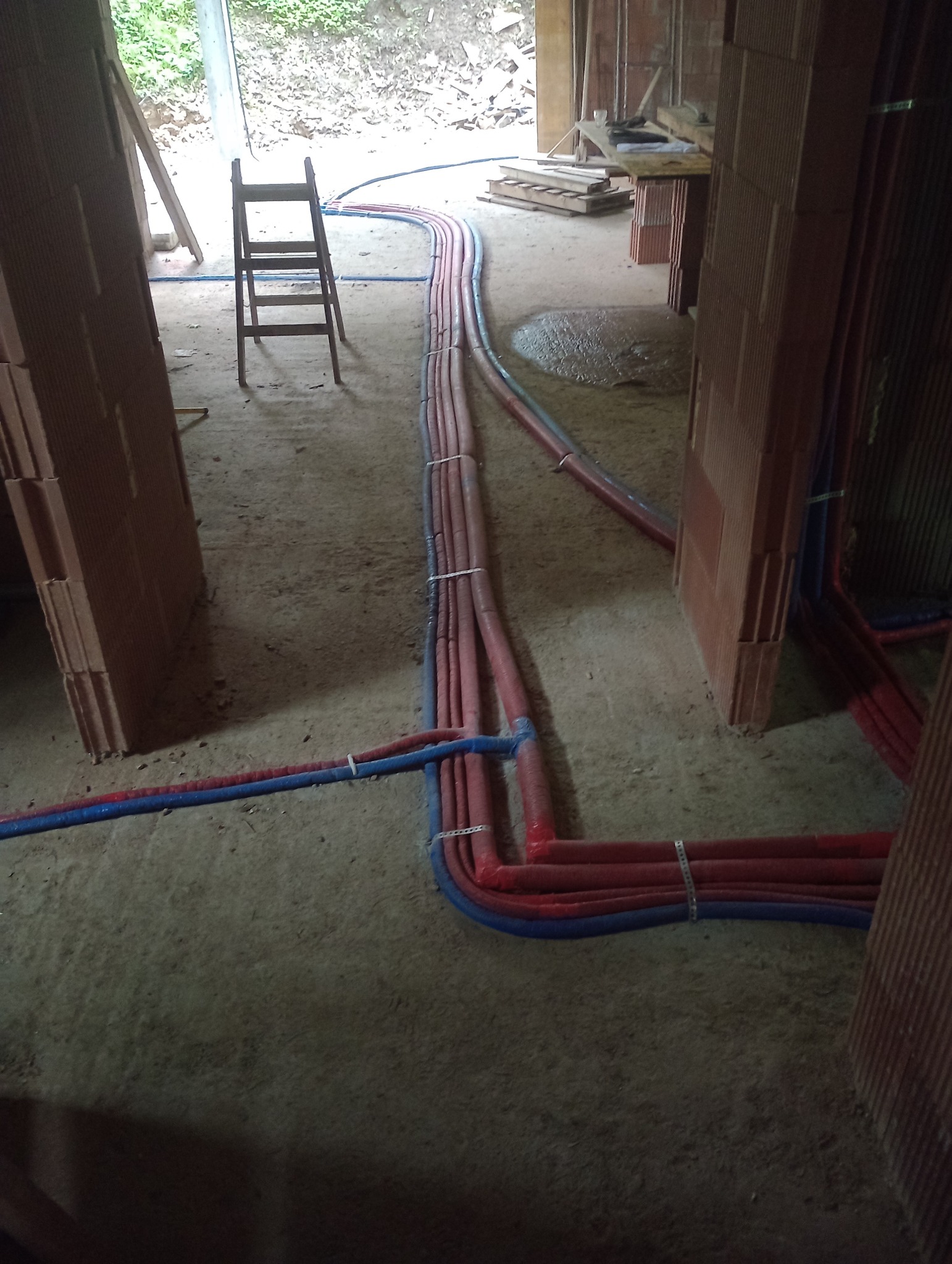 Rozprowadzenie rur instalacji wodnej i grzewczej w surowym stanie budynku z cegły, widoczna drabina i materiały budowlane w tle.