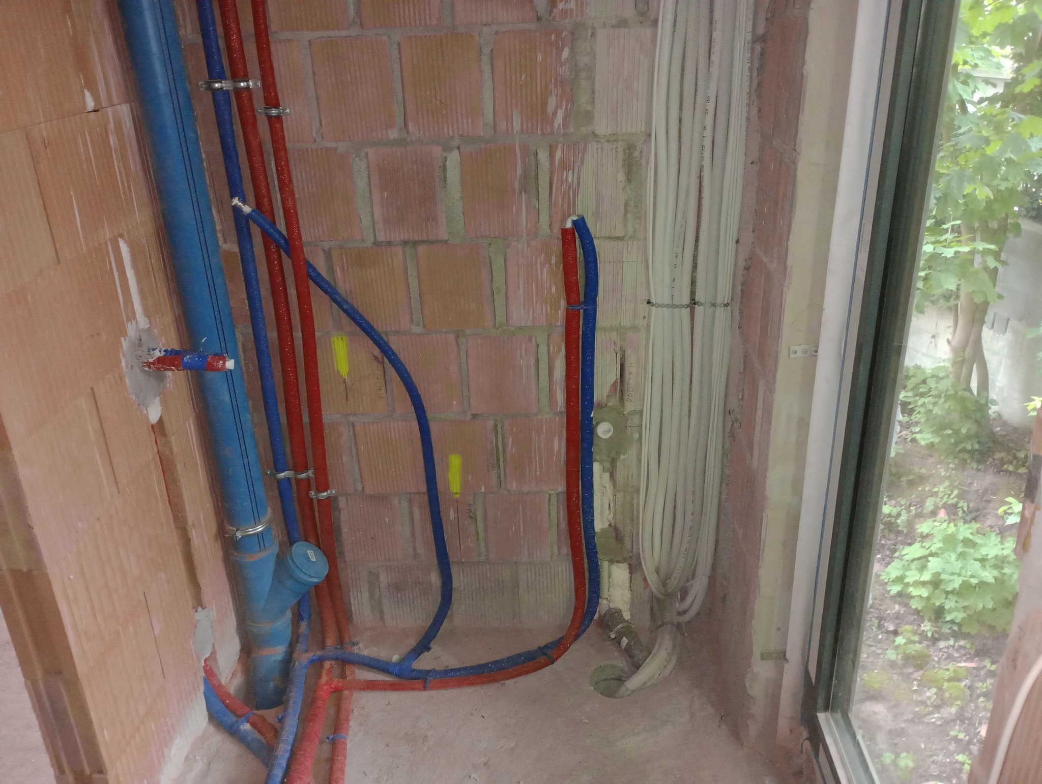 Instalacja wodno-kanalizacyjna w surowym stanie: czerwone i niebieskie rury prowadzone wzdłuż ceglanej ściany, widoczne mocowania i fragment okna z widokiem na zieleń.
