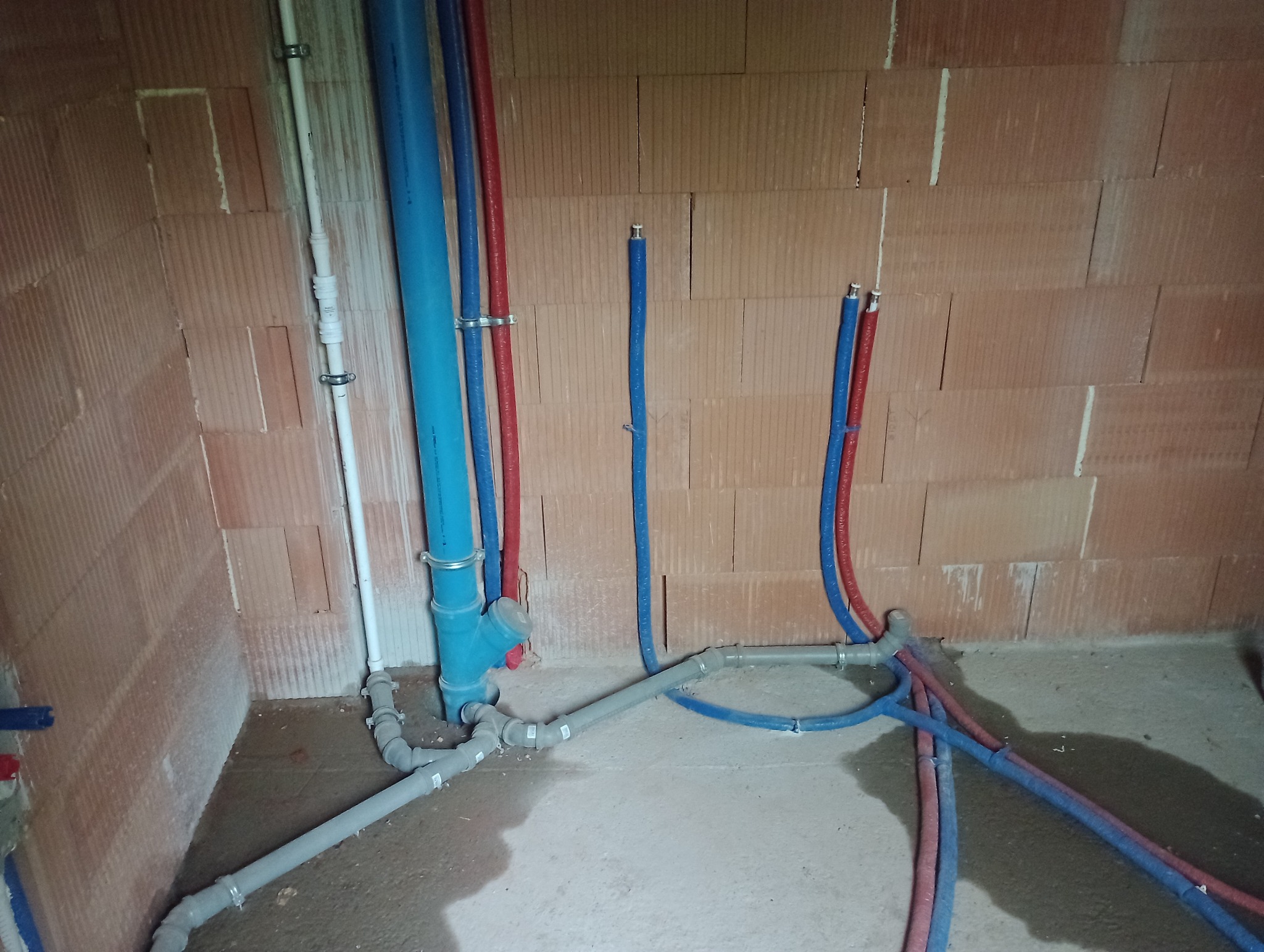 Wewnętrzne instalacje wodno-kanalizacyjne w surowym stanie, widoczne rury z tworzywa w kolorach niebieskim i czerwonym przymocowane do ściany z pustaków ceramicznych, szare rury kanalizacyjne...