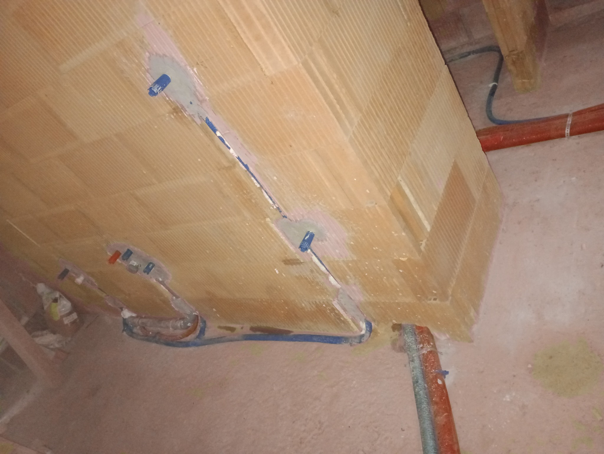 Instalacja ogrzewania podłogowego w bloczkach ceramicznych, widoczne niebieskie rury PEX zalane w zaprawie, częściowo zamontowane w ścianie.